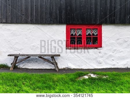 White house wall with red window on old faroese house. Wooden bench on green grass. Faroe islands, Denmark