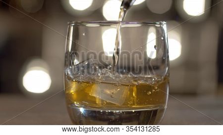 Close Up Pouring Whiskey Into Drinking Glass With Ice Cubes. Bar Counter On The Background Of Blurry