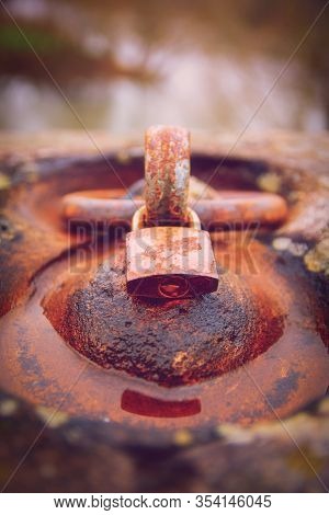 Close Up Of A Rusty Padlock Locked Around An Aged Rusty Iron Ring In A Puddle Of Water. Time And Agi