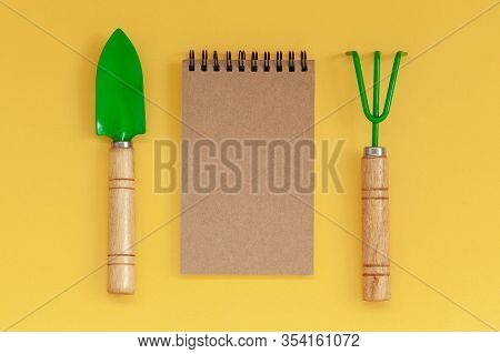 Wooden Handled Garden Hand Trowel And Hand Fork And Empty Noteboook On Yellow Background