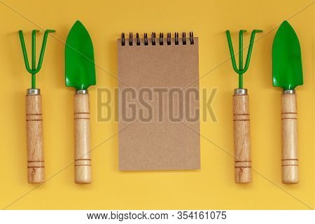 Wooden Handled Garden Hand Trowel And Hand Fork And Empty Noteboook On Yellow Background