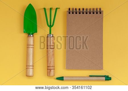 Wooden Handled Garden Hand Trowel And Hand Fork And Empty Noteboook On Yellow Background