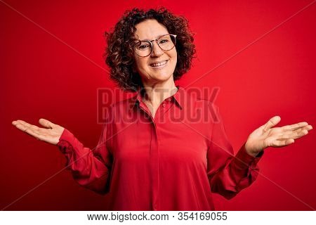 Middle age beautiful curly hair woman wearing casual shirt and glasses over red background smiling showing both hands open palms, presenting and advertising comparison and balance