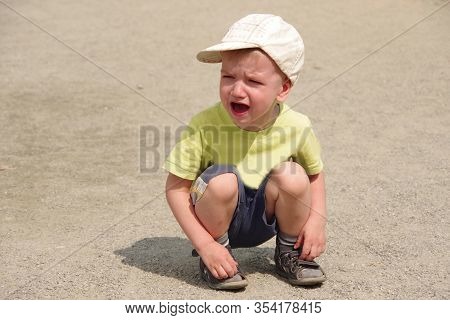 Caucasian Boy In A Yellow T-shirt, Dark Shorts, A Light Cap Sits On The Road And Cries. Family Value