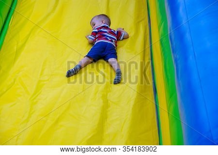 A Boy In A Striped T-shirt, Blue Shorts Rolls Off An Inflatable Slide. Happy Childhood. Outdoor Game