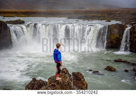 Young Hiker Standing At The Godafoss Waterfall In Iceland. Godafoss Means The Waterfall Of The Gods 