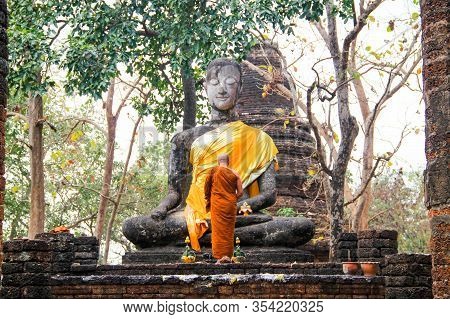 A Buddhistic Monk In Orange Dress Is Praying In Front Af An Ancient Statue