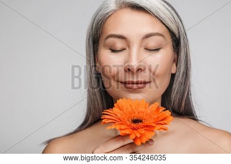Beauty portrait of an attractive sensual mature topless woman with long gray hair standing isolated over gray background, posing with herbera flower