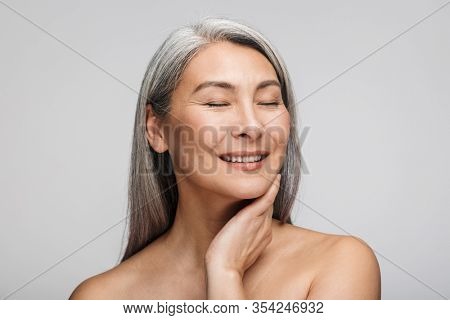 Beauty portrait of an attractive smiling mature topless woman with long gray hair standing isolated over gray background, ees closed