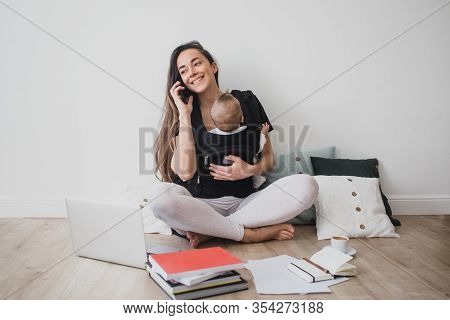 Young Freelance Mother Having Her Baby In Ergo Backpack And Working At Home Using Laptop And Smart P