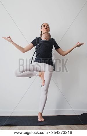 Young Mother Doing Yoga Exercises And Meditation With Her Little Baby In Ergo Backpack.