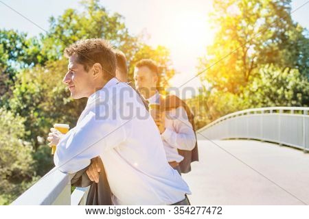 Portrait of mature thoughtful attractive businessman drinking coffee while standing on the bridge with colleagues with yellow sunshine in background