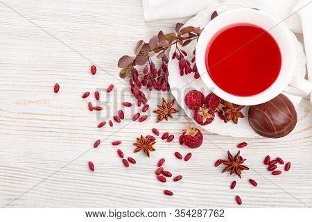 Healthy fruit tea with anise, barberries and raspberries in white porcelain cup and branch of berberry on light wooden background. Top view
