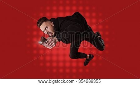 Handsome man in black clothes in motion in flight with a gun. Super agent on a mission. Criminal world, counterterrorism. Studio shot on a red background.