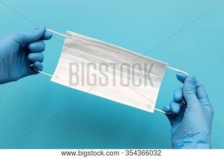 Hands Of Medic Wearing Blue Latex Gloves Holding Surgical Ear-loop Mask On Blue Background. Protecti