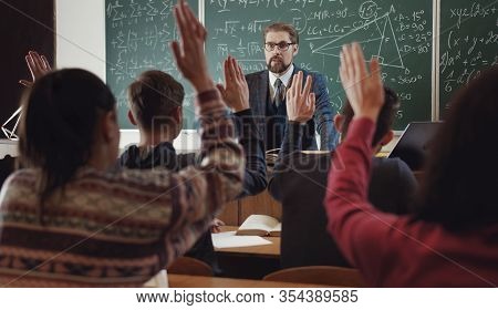 Group Of Students All Raising Hands To Answer Teachers Question, Focus On Tutor, View From Behind