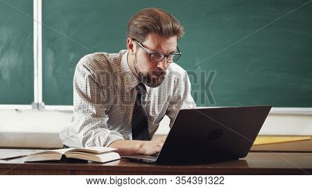 Serious Mature Bearded Teacher Wearing Shirt, Tie, And Spectacles Working On Laptop In Classroom