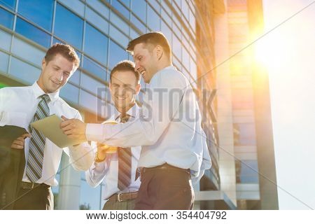 Mature attractive businessman showing and discussing plans over digital tablet with colleagues against office building