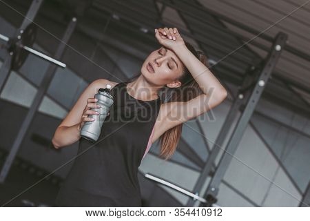 Exhausted Attractive Sports Woman Holding Bottle Of Water In Hand And Relaxing While Standing In Dar