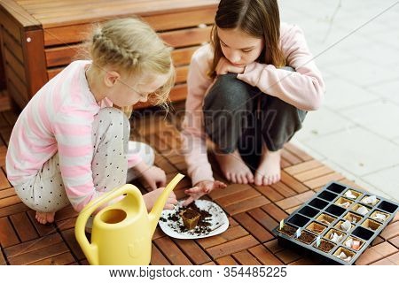 Two Cute Young Sisters Planting Flowers And Vegetables In Small Biodegradable Seedling Pots. Childre