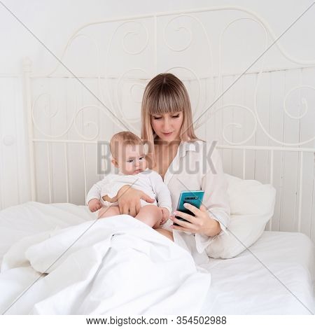 A Mum Blonde With Long Hair In Bed With A Baby In Her Arms Looks At The Phone, Sitting In Social Net