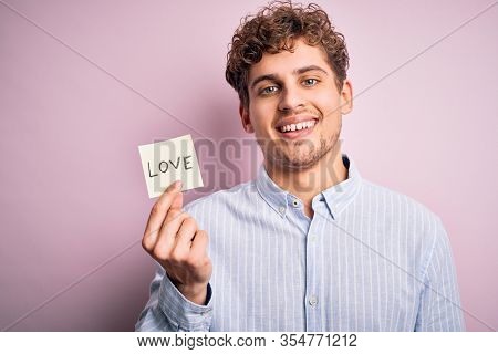 Young blond romantic man with curly hair holdiing paper with love message for valentines day with a happy face standing and smiling with a confident smile showing teeth