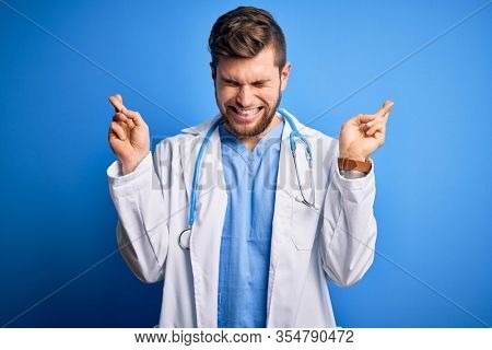 Young blond doctor man with beard and blue eyes wearing white coat and stethoscope gesturing finger crossed smiling with hope and eyes closed. Luck and superstitious concept.