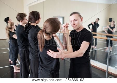 Young instructor of modern ballet dancing cource standing by one of his students while helping her with exercise at lesson