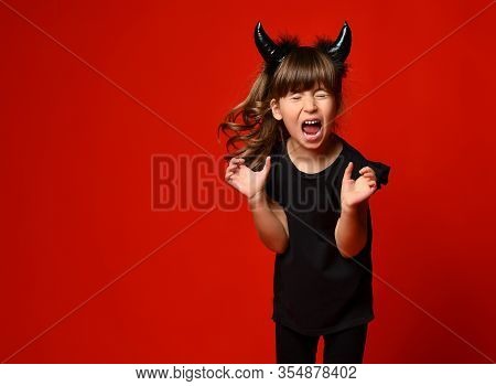 Little Brunette Kid With Devil Horns, Ponytail, In Black Blouse And Leggings. She Is Screaming With 