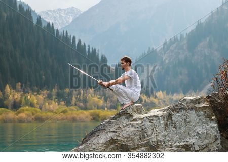 A Barefoot Man Sits On A Stone And Holds A Sword In His Hands. Preparing For The Battle. Man Health 