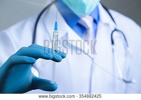 Doctor In Gauze Mask And Medical Robe Holding Syringe In His Blue Latex Glove Dressed Hand. Health A