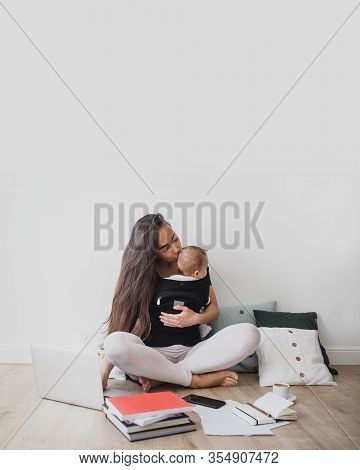 Young Freelance Mother Having Her Baby In Ergo Backpack And Working At Home Using Laptop And Smart P