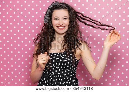 Kinky Brunnete Girl In Nice Black Polka-dot Dress Smiling Pleasantly, Wrapping Lock Of Hair Around H