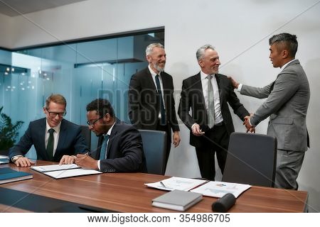 Two Happy Mature Businessmen In Classic Wear Shaking Hands While Having A Multi-ethnic Meeting In Th