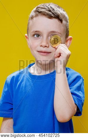 Little Happy Preschool Boy In A Blue T-shirt Holding Bitcoin In Front Of His Eyes On Yellow Backgrou