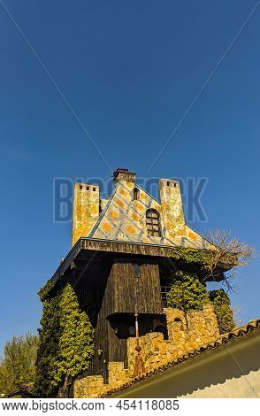 A Small Treehouse With A Sloping Roof And A Pair Of Chimneys And Small Windows Behind A Fence