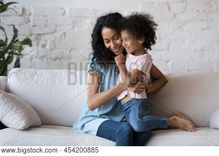 Excited Cheerful African Mom Tickling Happy Daughter Girl On Couch