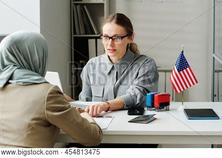 Modern Caucasian Consular Officer Sitting At Desk In Front Of Muslim Woman Explaining Her How To Fil