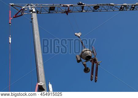 A Round Grey Concrete Mixer And A Red Movable Carriage Trolley Are Suspended From The Boom Hook Of A