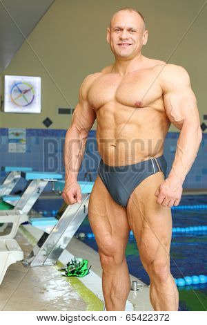 Happy sunburnt bodybuilder stands near indoor pool before swimming in gym hall