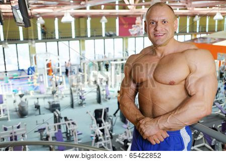 Smiling shirtless sunburnt bodybuilder stands in big modern gym hall