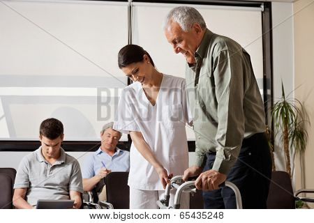 Senior man being assisted by female nurse to walk the Zimmer frame with people sitting in background