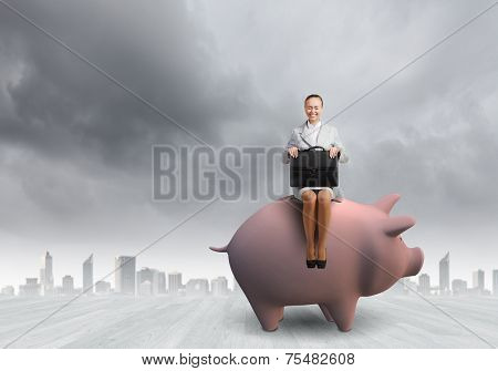 Young businesswoman with suitcase in hand sitting on piggybank