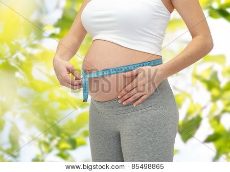 pregnancy, motherhood, control, people and expectation concept - close up of happy pregnant woman measuring her bare tummy over green tree leavers background