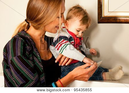 Young beautiful mother and her cute  little boy are looking and playing with a smartfone indoors. Copy space frame.
