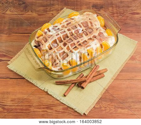 Sweet pancakes filled with cottage cheese with sour cream and sprinkled with cinnamon powder and mandarin orange segments in the rectangular glass roasting pan and cinnamon sticks on cloth napkin on old wooden surface