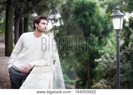 Dreamy young handsome man waiting for the first meeting. Latin lover. A beautiful and young Italian boy is leaning on a wall outdoors in a park. He looks at a lamppost in the town.He is waiting for his girlfriend? Concept of love, waiting, passion.