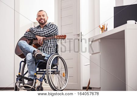 Enjoying every moment of it. Attractive smiling young man looking delighted while posing for a photographer and sitting on a wheelchair in his apartment