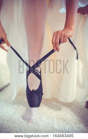 Closeup legs of young ballerina who puts on pointe shoes at white class room background. Ballet practice on training. Beautiful slim graceful feet of ballet dancer unrecognizable female.