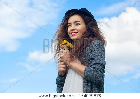 Attractive, Young Girl In Jeans And A Black Hat Holding A Blooming Dandelions, Smiling On The Backgr